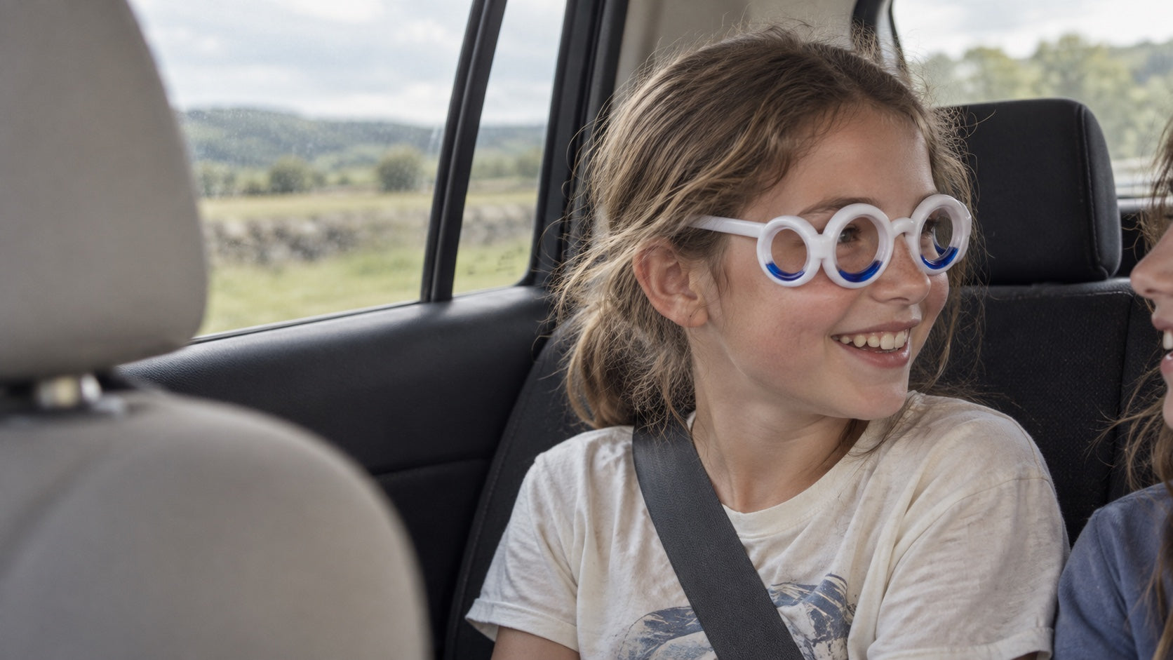A child wearing Ollaway and a seatbelt smiles toward a sibling on a car journey